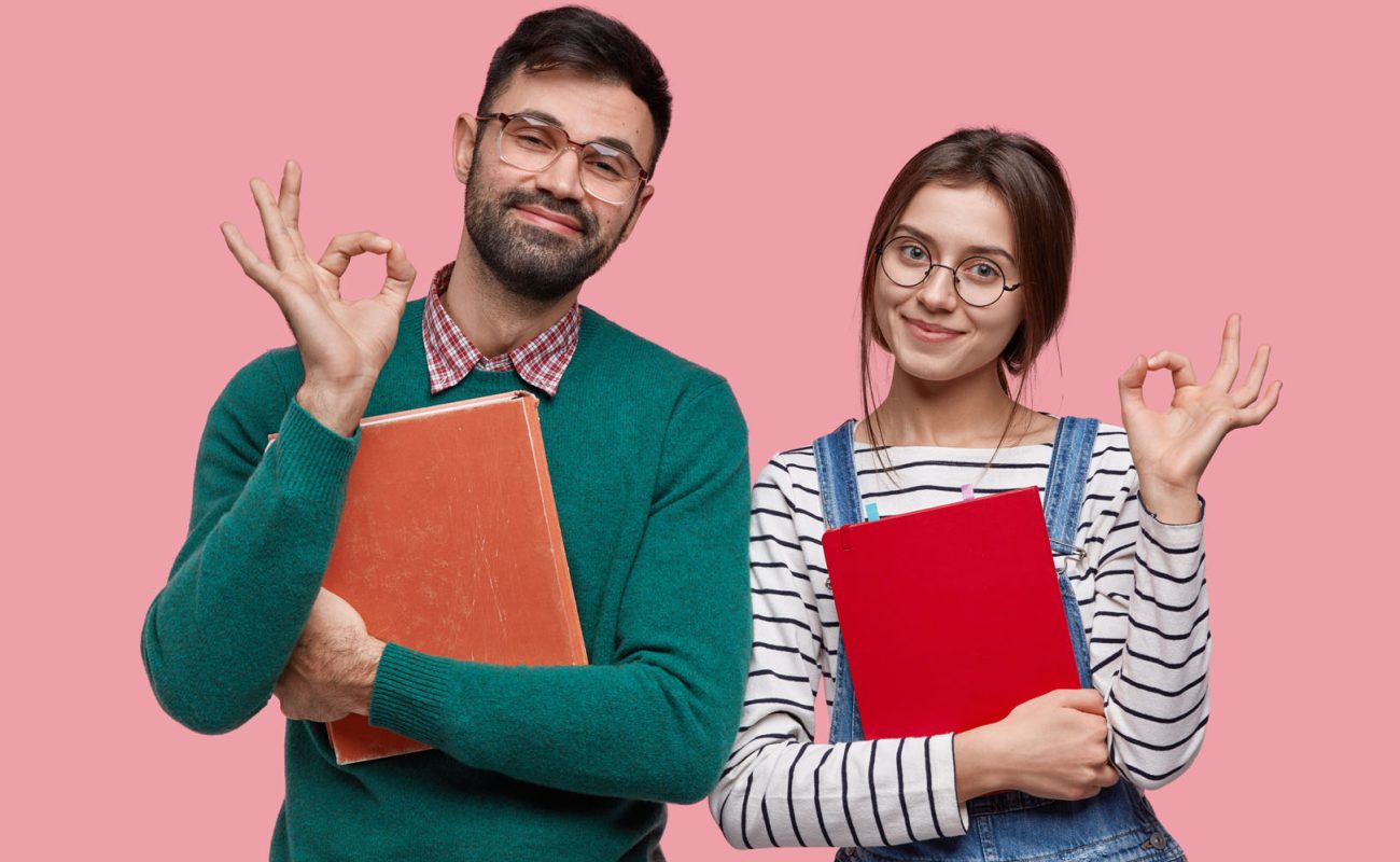 Photo of handsome male student and his female groupmate demonstrates okay gesture, agree with something, hold book and notebook closely, isolated over pink background. Everything is perfect!